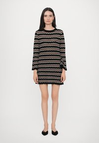 Robe en tricot avec un motif en zigzag noir, blanc et rose clair. Dotée de manches longues et d'un ourlet en dentelle. Portée avec des chaussures noires.