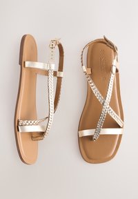Sandali bassi con cinturino intrecciato dorato, dettagli metallici e suola marrone chiaro dalla texture liscia. Chiusura con fibbia inclusa.