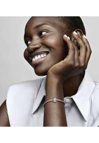 Pulsera de plata con un diseño texturizado, que presenta un cierre decorativo. Modelo con el cabello corto reposa la mano en la barbilla, vistiendo una blusa gris claro.