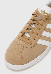 Baskets en suede beige avec rayures blanches, bout rond, design à lacets plats et semelle texturée. Présentent un accent logo doré sur la languette.