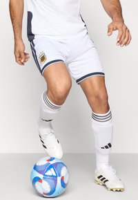 Λευκά σορτς ποδοσφαίρου με μαύρες ρίγες και έμβλημα, σε συνδυασμό με λευκές κάλτσες και μαύρα-χρυσά παπούτσια. Μια μπάλα ποδοσφαίρου με μπλε και κόκκινο σχέδιο είναι στα πόδια.
