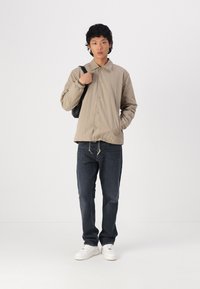 Chaqueta beige ligera con cuello de solapa, botones delanteros y puños elásticos. Combinada con jeans de mezclilla oscura y zapatillas blancas.
