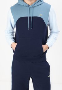 Mörkblå och ljusblå färgblockerad hoodie med ficka, dragskohet och Lacoste-logotyp. Mjuk, texturerad bomullsmaterialblandning.