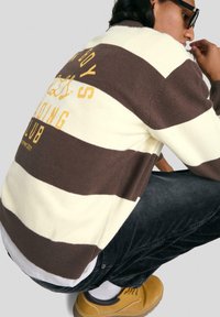 Csíkos pulóver krém és barna gyapjú keverékből, hátul sárga grafikával. Fekete kordbársony nadrággal és sárga cipőkkel párosítva.