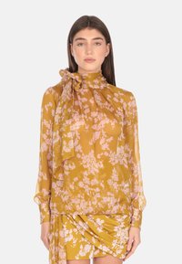 Blusa floreale gialla con maniche lunghe, tessuto morbido, colletto alto e dettaglio a fiocco al collo; presenta motivi floreali rosa su tutto.