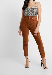 Pantaloni in suede marrone a vita alta, gamba affusolata, abbinati a una canotta con stampa pitonata nera e grigia e sandali neri con tacco.
