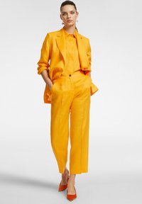Completo di lino giallo con giacca sartoriale e pantaloni larghi, abbinato a tacchi arancioni a punta. Tessuto testurizzato con vestibilità casual.