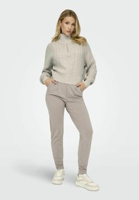 Helles graues gestricktes Sweatshirt mit hohem Kragen und Kabelstrickmuster, kombiniert mit beigen schmal geschnittenen Hosen und weißen Turnschuhen.