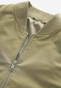 Gros plan d'une veste bomber beige avec col côtelé et fermeture éclair argentée partiellement ouverte, montrant une texture de tissu lisse.
