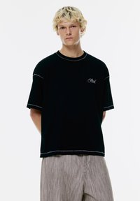 Czarny t-shirt z krótkim rękawem z kontrastowymi białymi szwami i haftowanym tekstem na piersi, w połączeniu z jasnoszarymi spodniami w paski.