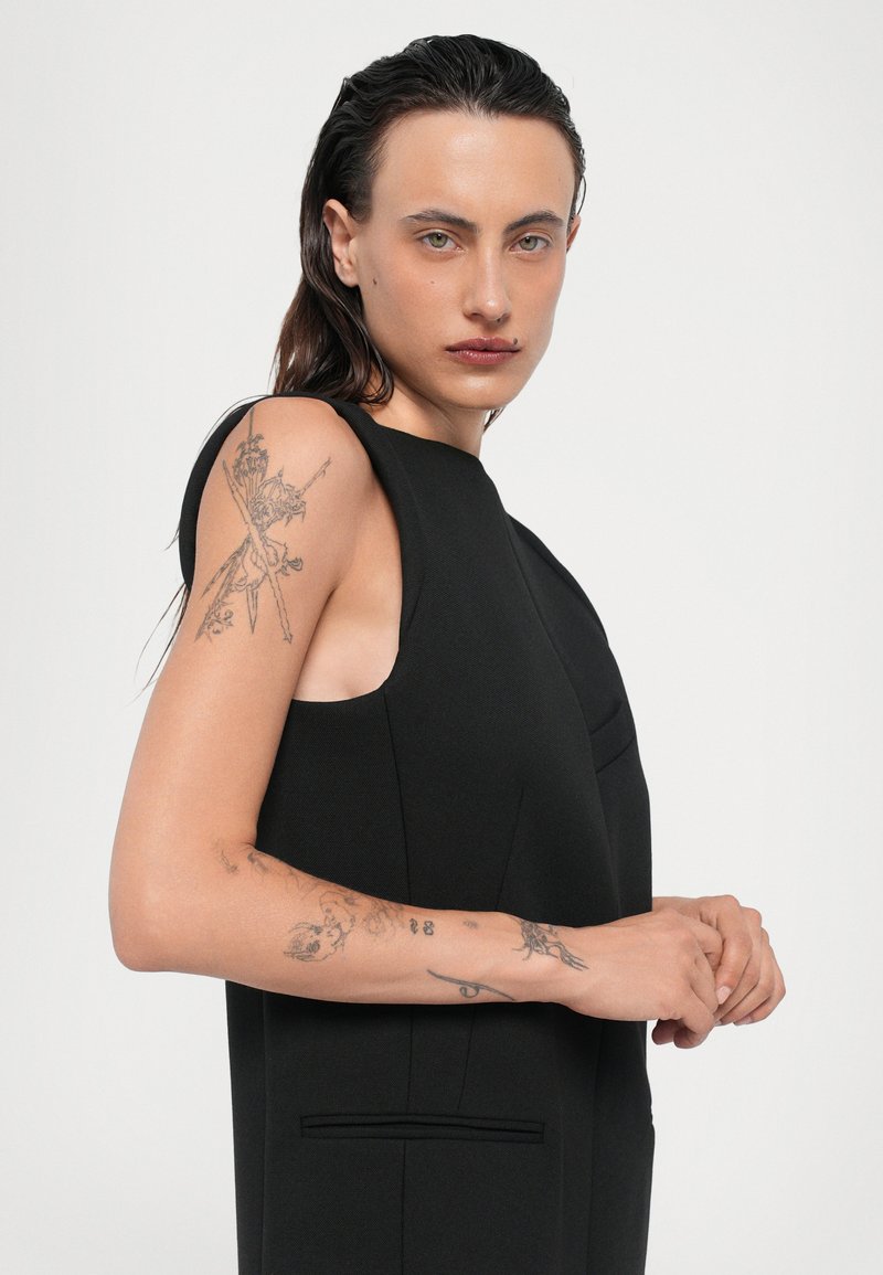 Svart ärmlös klänning med en figursydd design och subtila detaljer. Har texturerat tyg och sidofickor, med synliga arm-tatueringar.