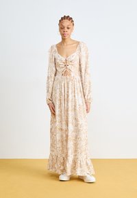 Robe longue à motifs floraux en beige clair avec des accents crème, présentant un décolleté profond, une taille froncée et des manches longues, assortie de chaussures blanches.