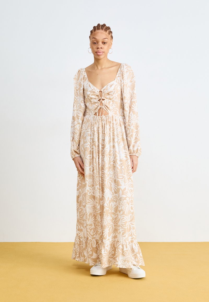 Robe longue à motifs floraux en beige clair avec des accents crème, présentant un décolleté profond, une taille froncée et des manches longues, assortie de chaussures blanches.