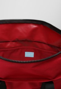 Interno di una borsa di stoffa rossa con una texture liscia e un piccolo tag blu con l'etichetta "Calvin Klein Jeans". Cerniera nera lungo l'apertura.