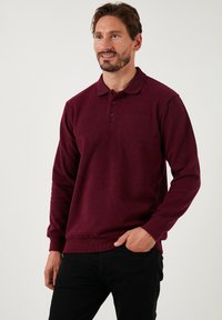 Burgunderfarbener Langarm-Polohemd aus strukturiertem Stoff mit einer Drei-Knopf-Leiste und gerippten Bündchen. Kombiniert mit dunklen Jeans.