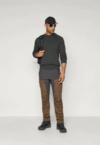 Mørkegrå strikkegenser, grå og brune cargo-lignende bukser med lommer, svarte støvler, svart caps og solbriller, med en ryggsekk på den ene skulderen.