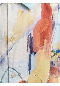 Lähikuva paidan kankaasta, jossa on abstrakteja akvarellikuvioita sinisessä, oranssissa, keltaisessa ja punaisessa, näkyen nappeja ja osa kaulusta.