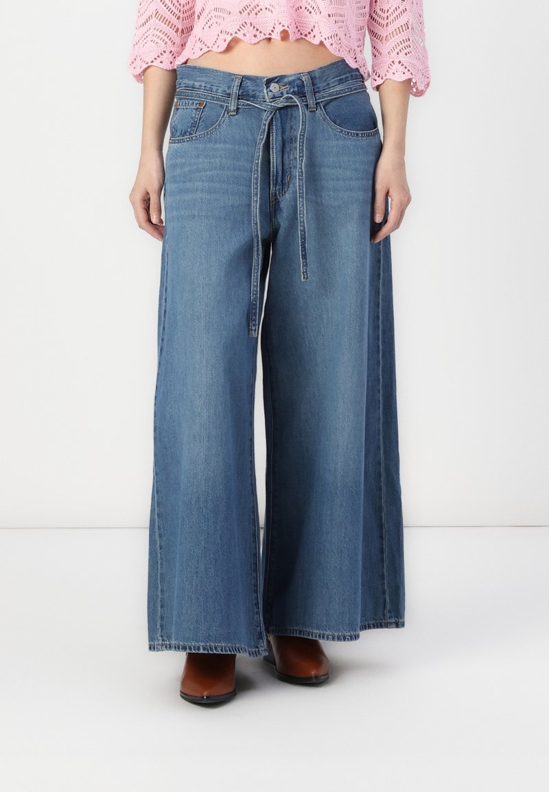 Femme portant un jean bleu taille haute à jambes larges avec une ceinture nouée en denim, associé à des chaussures marron et un top court rose en crochet.