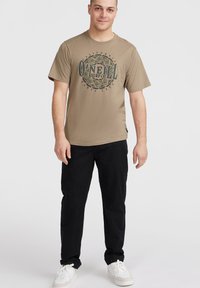 Camiseta de manga corta color caqui con un gráfico circular azul y verde que lleva el texto "O'NEILL SURF CO" en el frente. Pantalones negros y zapatillas blancas.