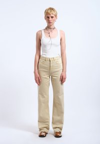 Jong persoon met kort blond haar, een wit tanktop, beige hoog getailleerde spijkerbroek, gelaagde kettingen en bruine schoenen met patroon, staand tegen een witte achtergrond.