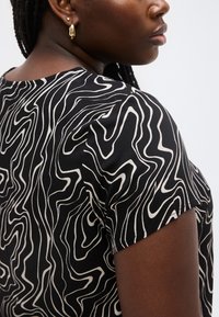 Άτομο με σκουρόχρωμο δέρμα που φοράει κοντομάνικη μπλούζα με ασπρόμαυρο αφηρημένο μοτίβο, χρυσά και μαργαριταρένια σκουλαρίκια, με πλεγμένα μαλλιά, γυρισμένο στο πλάι.