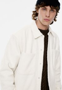 Valkoinen fleece-takki kauluksella, kahdella rintataskulla ja napillaan suljettava. Päällä ruskea palmikkoneulepaita.