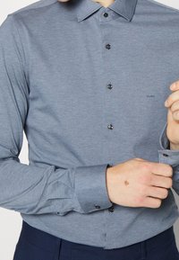Langsleeve shirt van lichtblauw-grijs stof, met een button-down kraag, zwarte knopen en subtiele logo borduursel op de borst.