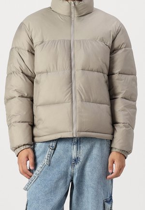 Homme portant une veste matelassée beige clair avec fermeture éclair à l'avant et poignets élastiques, associée à un jean bleu denim ample avec détail de sangle.