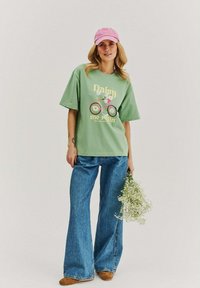 Young woman wearing green graphic t-shirt, blue wide-leg jeans, pink cap, holding a bouquet of small white flowers.