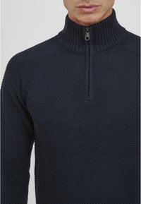 Marineblaues Zip-Up-Pullover mit hohem geripptem Kragen, der ein strukturiertes Strickmuster aufweist und einen Reißverschluss in Viertellänge am Hals hat.
