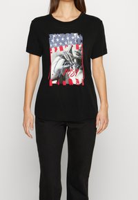 Sort bomulds T-shirt med et grafisk print, der viser et vintage billede, farverne fra det amerikanske flag og tekst der siger "1989" i rød og blå.