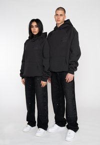 Black hoodies with kangaroo pockets and embroidered logo, paired with loose dark jeans and white sneakers, displayed on two models.