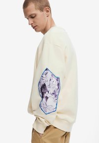 Crème kleurige sweatshirt met een blauw gestikte, grafische patch op de linkermouw met illustraties van cartoonfiguren. Ontspannen, oversized pasvorm.