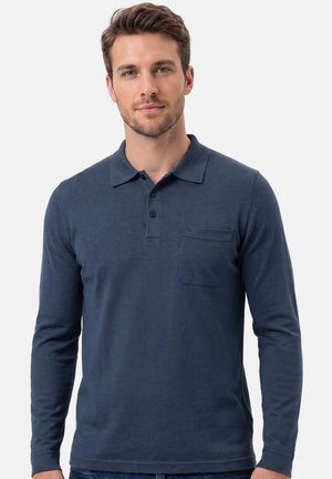 Mężczyzna ubrany w dopasowaną, granatową koszulkę polo z długim rękawem, kołnierzykiem, trzema guzikami i kieszenią na piersi, stojący na jednolitym tle.