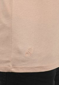 Lys beige t-skjorte med en glatt tekstur, med en liten brodert spiral-logo ved kanten. Korte ermer og en avslappet passform.