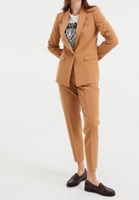 Tan suit with fitted blazer and tapered pants, paired with a graphic tee. Brown loafers complete the look. Smooth, lightweight fabric.