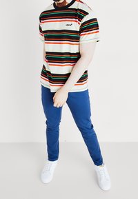 Pasiasta koszulka z krótkim rękawem w kolorach kremowym, czarnym, czerwonym i zielonym; logo na piersi. Połączona z niebieskimi spodniami i białymi trampkami.