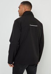 Schwarze wasserdichte Jacke mit hohem Kragen, ausgestattet mit einem reflektierenden Streifen über dem Rücken und strukturiertem Stoff. Slim Fit mit verdeckten Nähten.