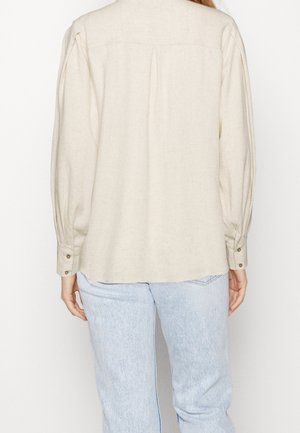 Kvinde iført en cremefarvet bluse med lange ærmer med knaplukning ved manchetterne og lyseblå jeans, vist bagfra mod en ensfarvet baggrund.