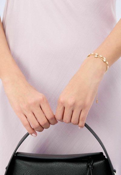 Brazalete de oro con acentos de estrellas y cuentas similares a perlas, llevado en la muñeca. Bolso negro texturizado sostenido con ambas manos. Vestido rosa claro visible.