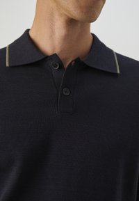 Polo bleu marine en tissu texturé, avec deux boutons noirs et une ligne d'accents vert pâle au col, soulignant un design classique.