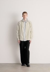 Casaco leve bege claro sobre uma camisa riscada, combinado com calças pretas e sapatos polidos, num modelo contra um fundo simples.