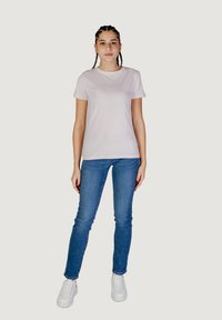 Lichtroze t-shirt met korte mouwen en het "GUESS" logo, gecombineerd met blauwe slim-fit jeans en witte sneakers, tegen een neutrale achtergrond.