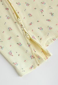 Gros plan sur un tissu côtelé jaune pâle avec de petites imprimés floraux roses et violets, présentant deux fermetures éclair beiges parallèles partiellement ouvertes.