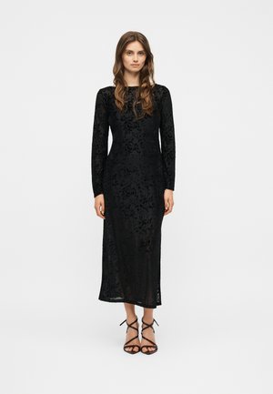 Γυναίκα με μακριά κυματιστά μαλλιά που φοράει ένα μαύρο, μακρυμάνικο, μακρύ μέχρι τον αστράγαλο βελούδινο φόρεμα με λουλούδια και μαύρα υψηλά πέδιλα με λουράκια.