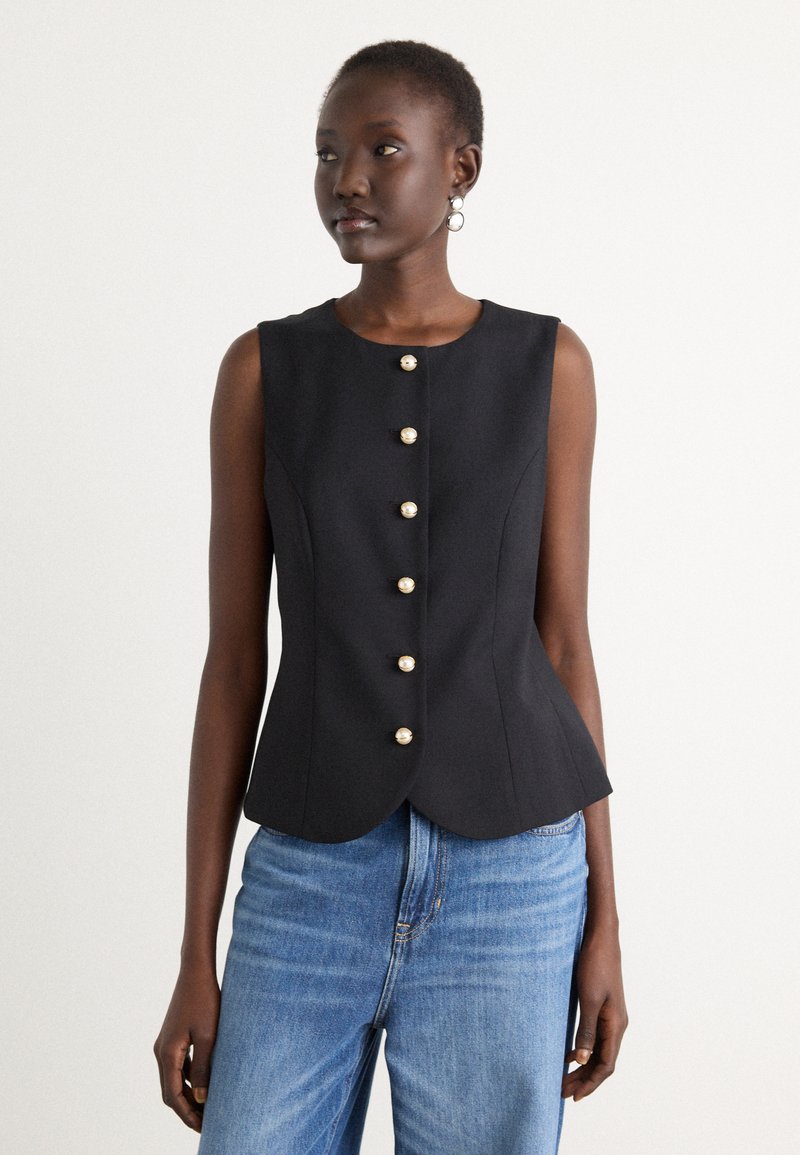 Svart ermeløs topp med tettsittende silhuett, rund hals, og åtte gullfargede knapper foran. Kombinert med lyseblå denim.