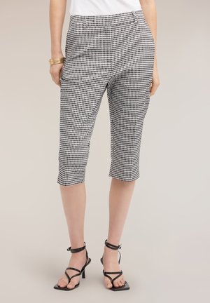 Vrouw met zwart-witte geruite knielange broek, wit topje, gouden armbanden en zwarte sandalen met hoge hak en bandjes, staand op een beige achtergrond.