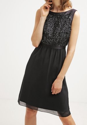 Hihaton musta polvipituinen mekko, jossa on teksturoitu, kiiltävä yläosa ja sileä helma, päällä mallin, joka seisoo valkoista taustaa vasten.