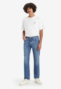 T-shirt blanche avec logo noir, jean en denim bleu avec une coupe droite, et baskets couleur crème. Le modèle se tient avec les mains dans les poches.