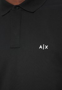 Camisa polo preta com colarinho abotoado e logótipo bordado em branco "A|X" no lado esquerdo do peito.